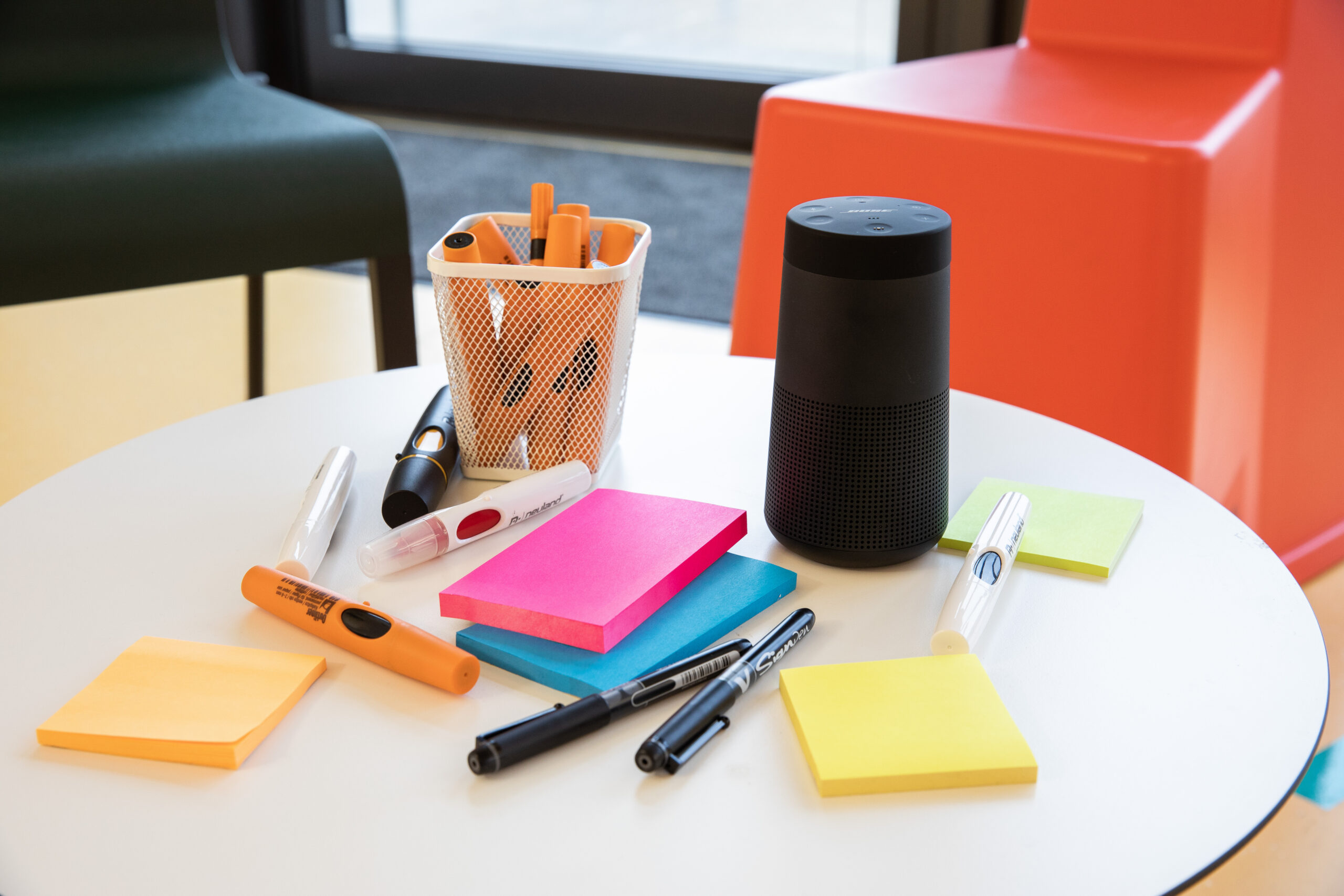 Kleiner, runder Tisch mit hochwertigen Arbeitsmatieral von Post-It, Neuland und schwarzer Bluetoothbox von Bose. Rechts sieht man einen schwarzen Stuhl und links einen roten Stuhl.