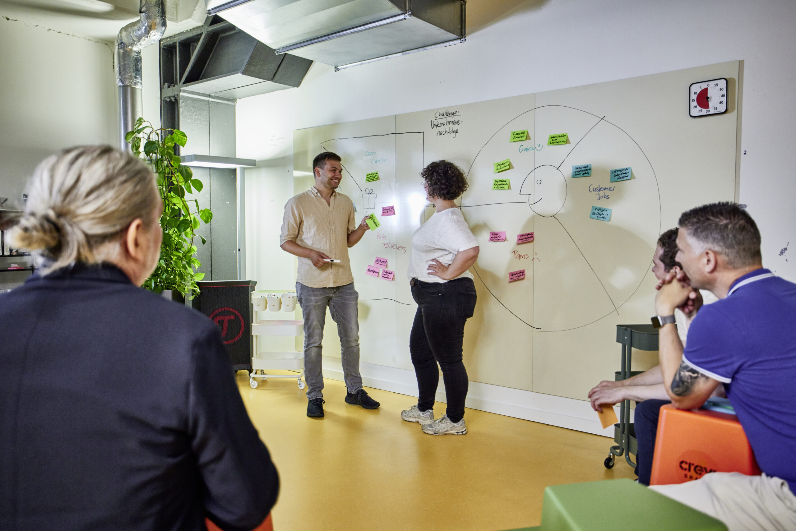 Zwei Personen stehen vor einem beschriebenen Whiteboard; eine Person zeigt der anderen lächelnd ein Post-it, mehrere Personen sitzen davor und schauen zu.