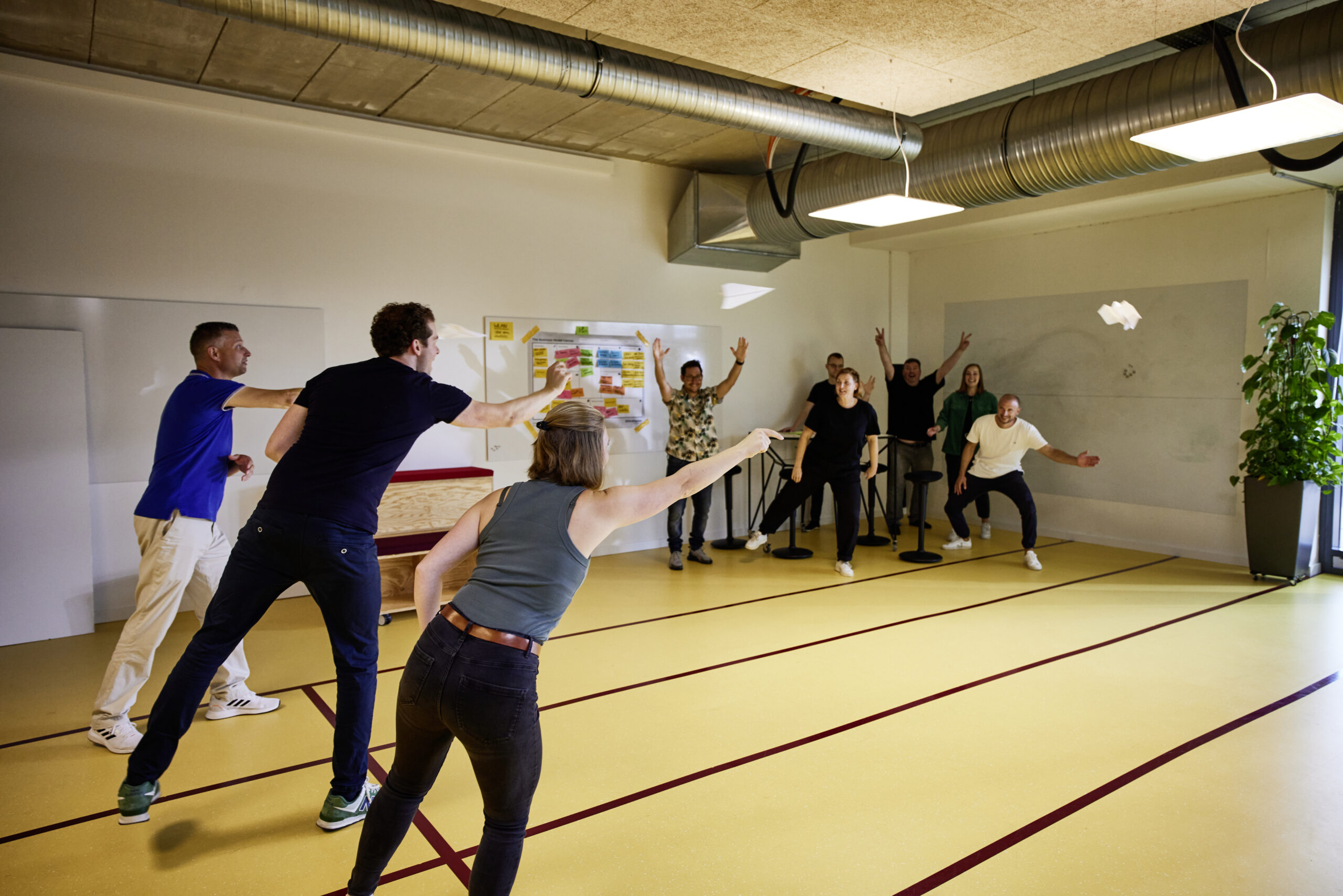 Teamevent in einem Workshopraum: Personen werfen Papierflieger, während im Hintergrund andere jubeln.