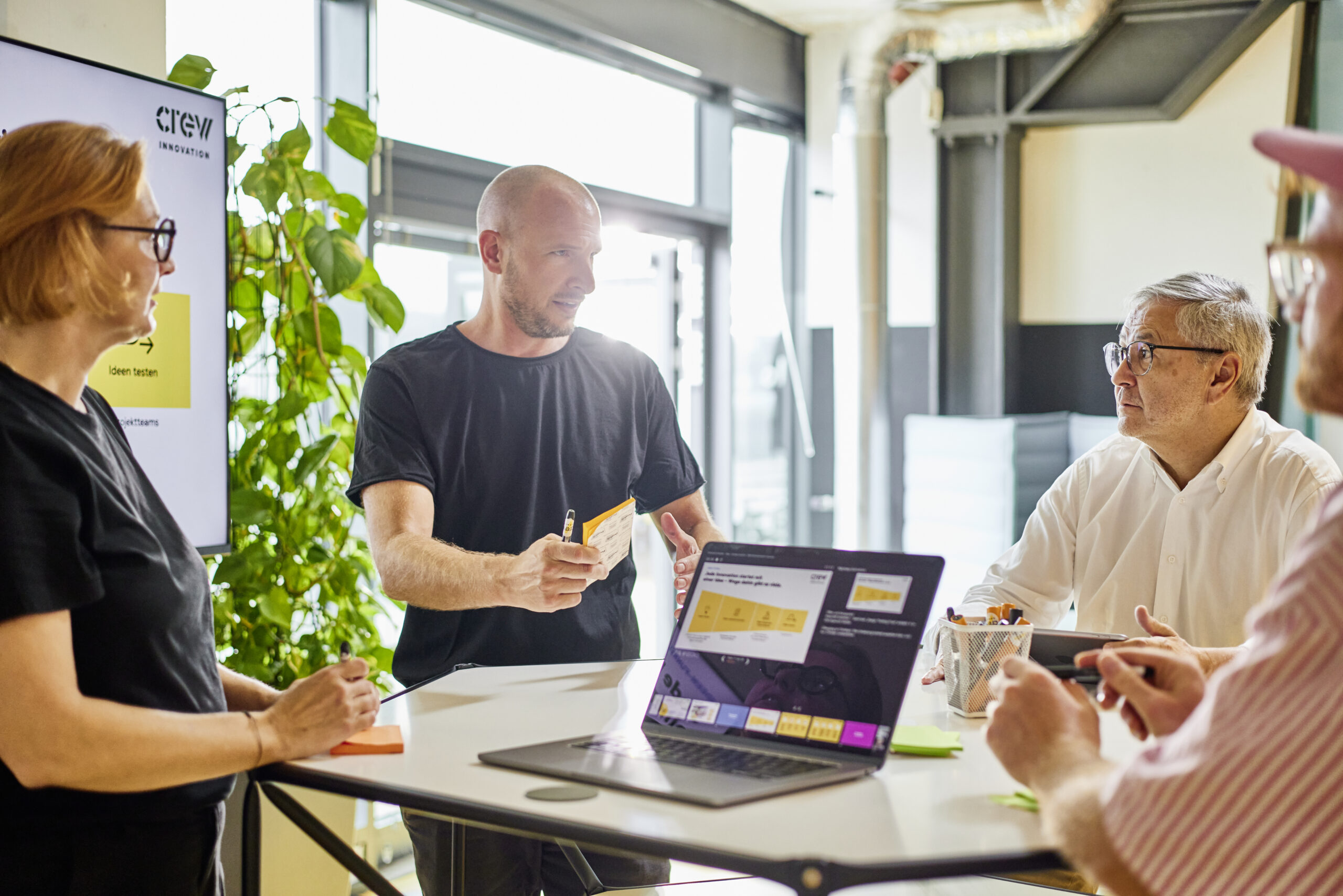 Personen stehen an einem Stehtisch und hören einer Person zu; im Hintergrund ist eine Präsentation geöffnet.