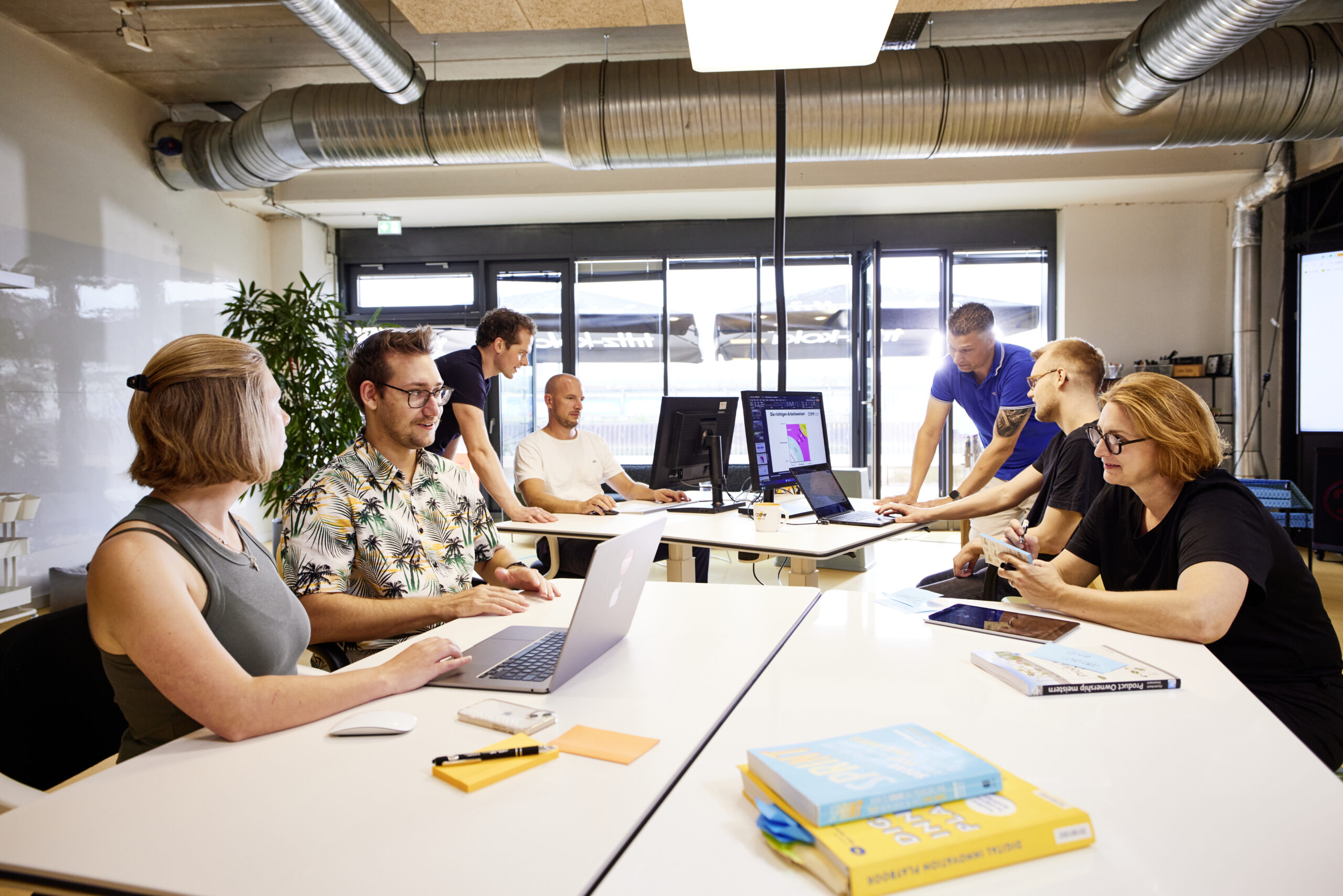 Mehrere Personen sitzen in Gruppen an zwei Tischen, arbeiten an Laptops und unterhalten sich miteinander.