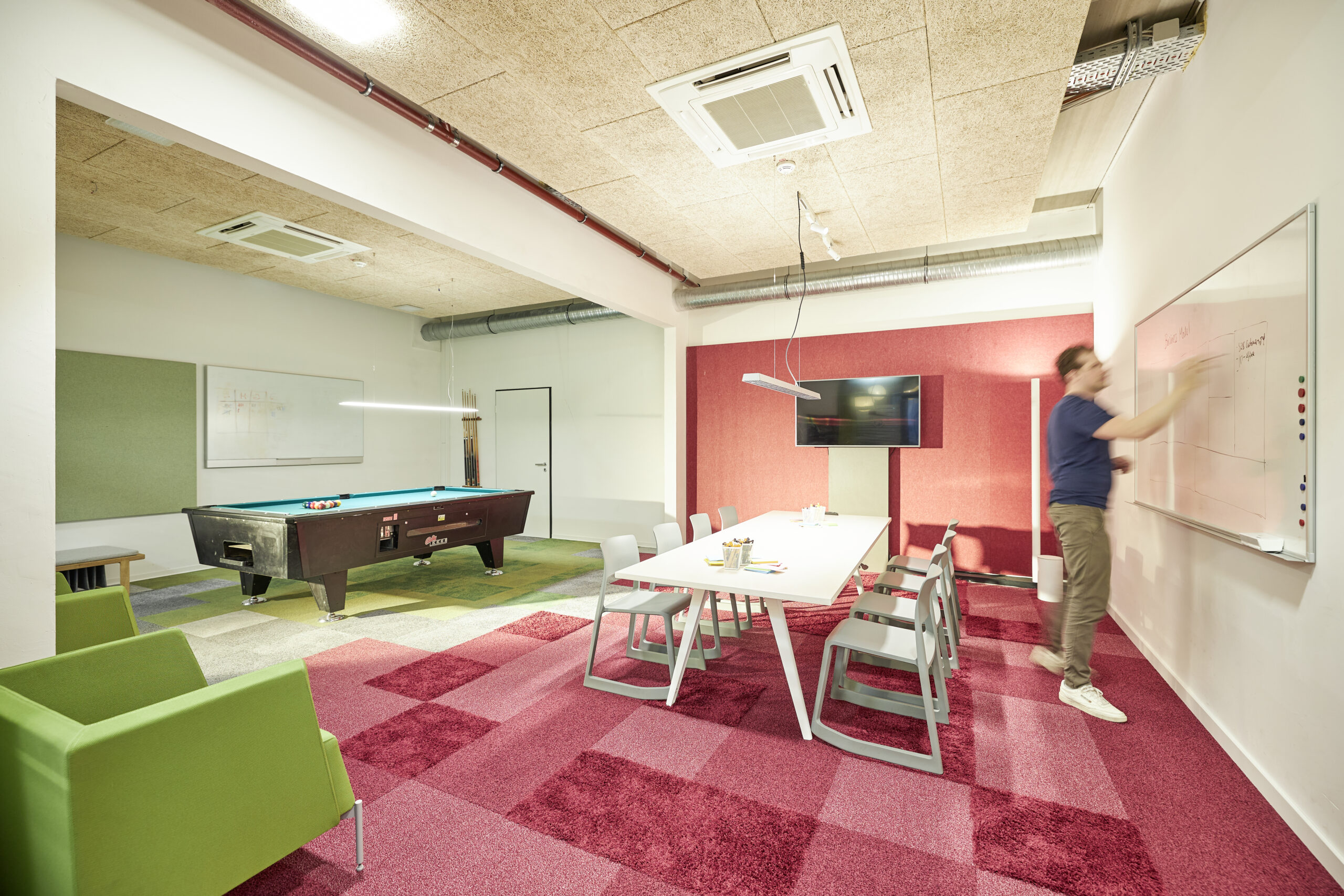 Moderner Konferenzraum mit Tisch, Fernseher und Billardtisch; eine Person schreibt etwas an ein Whiteboard.