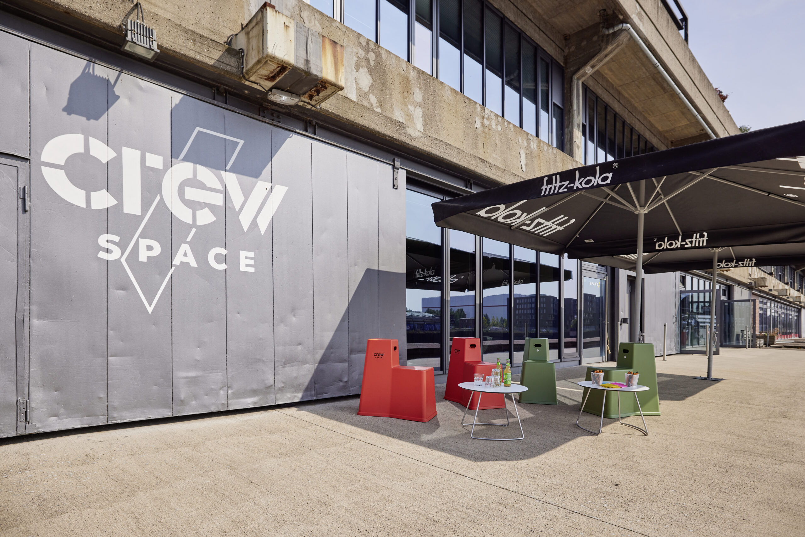 Sonnige Terrasse mit CREW Space Logo an der Wand und Sitzgelegenheiten unter großen Sonnenschirmen.
