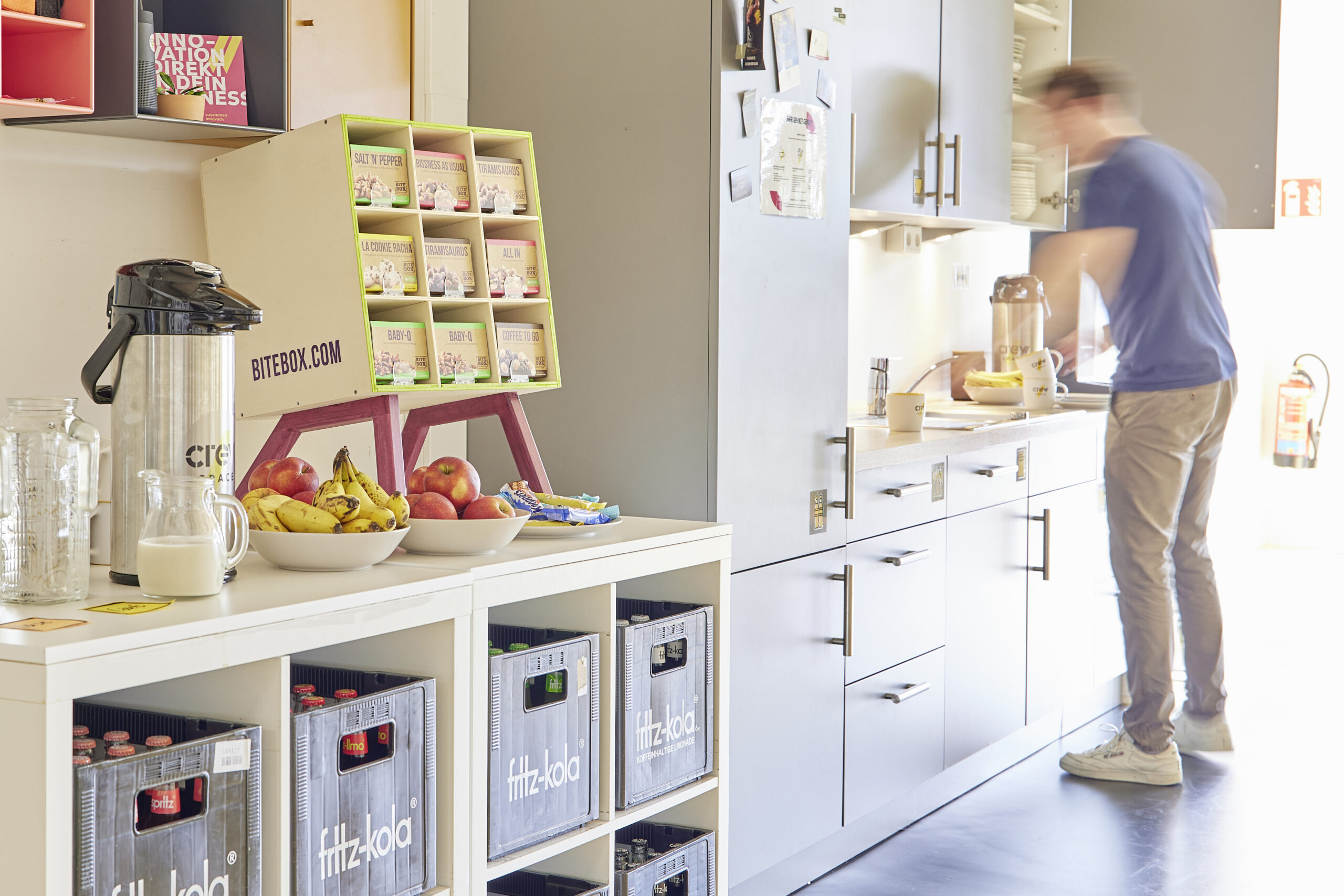 Küchenzeile und Gastro-Bereich im Workshopraum mit Obst, Kaffee und Snacks; eine Person steht am Waschbecken.