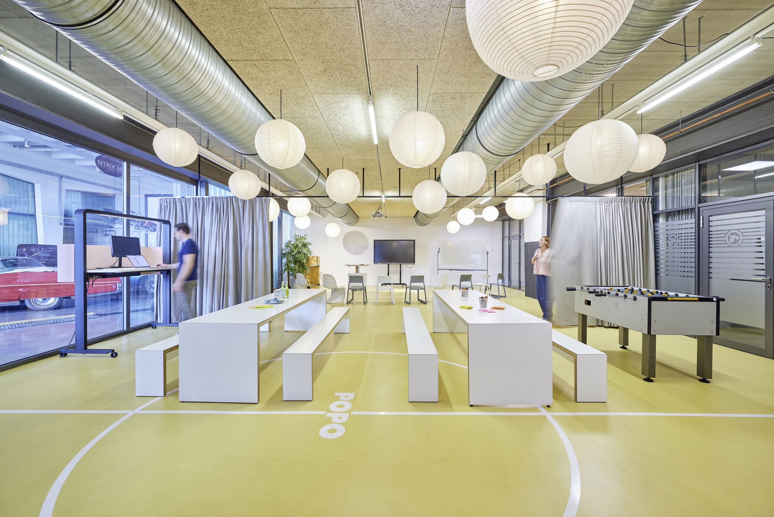 Moderner Workshopraum: Eine Person arbeitet am Laptop an einem Stehtisch, während eine andere den Vorhang schließt.